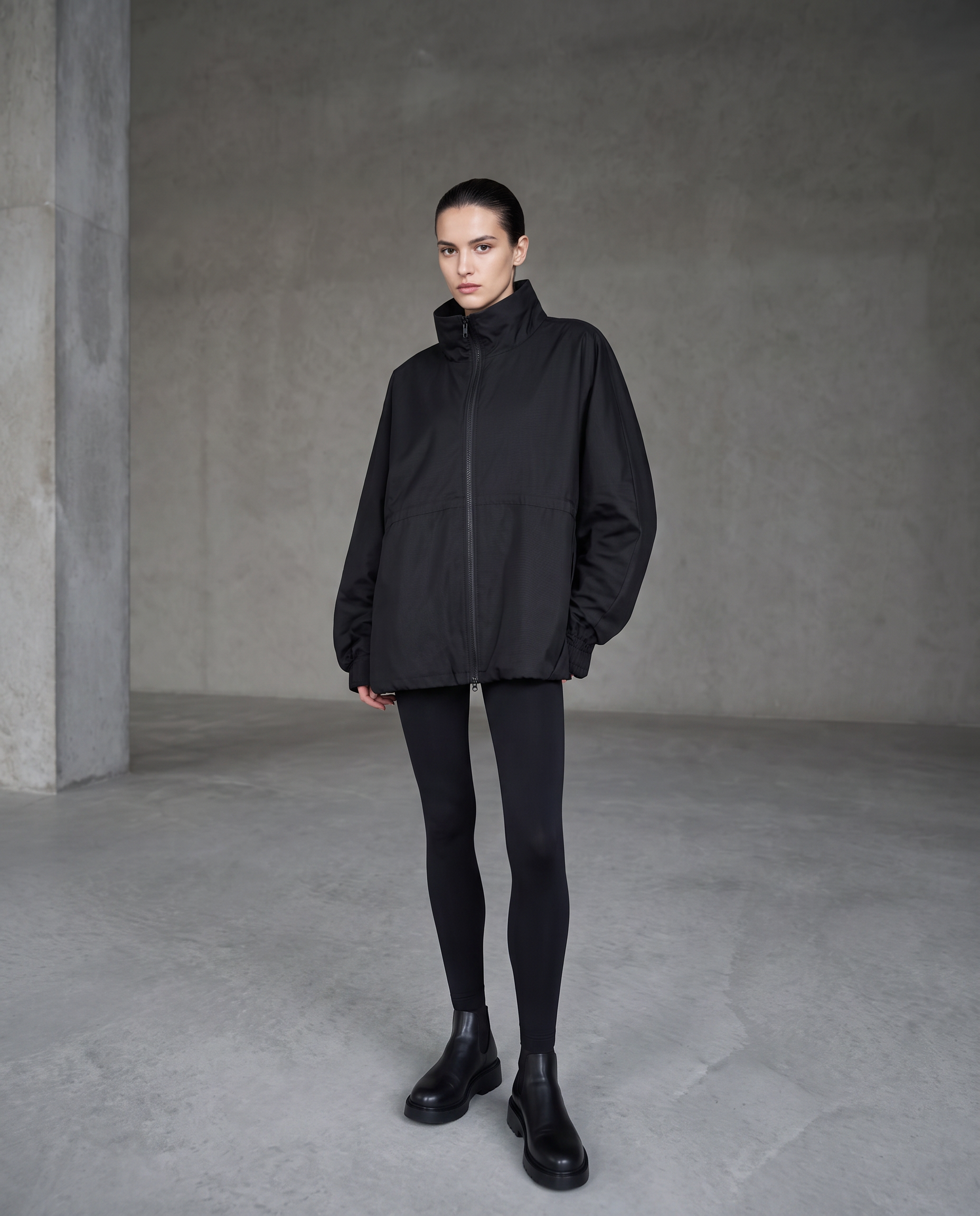 Model wearing an oversized black technical jacket and black leggings in a concrete space